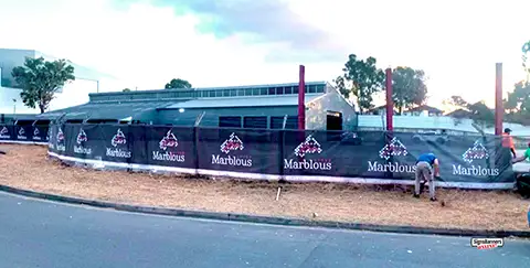 Shows a work crew unwinding the last section of the printed roll almost completing the fence project. Its a creative design of mesh banner on fence. The fence is typical size. The banner is 1600 millimetres high by 50 metres long and wraps around the fence curvature nicely.