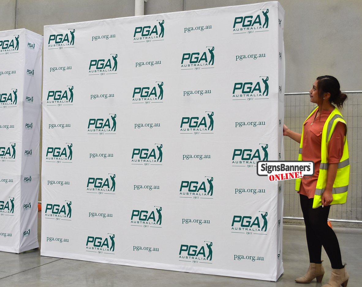 Front view of a white PGA Australia pop-up media wall with repeating logos, woman in hi-vis vest standing at the side.