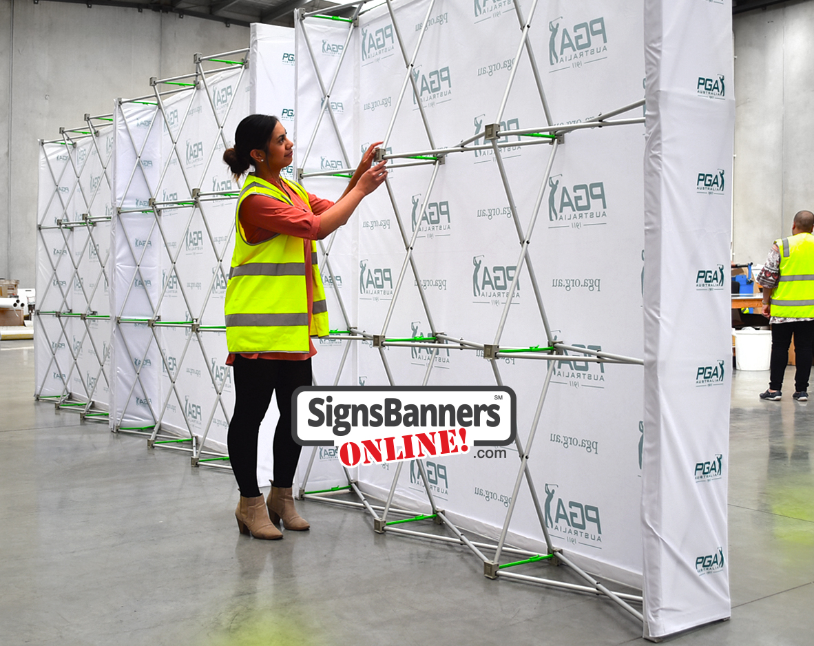 Woman in hi-vis vest assembling the aluminium frame behind a row of white PGA Australia pop-up media walls.