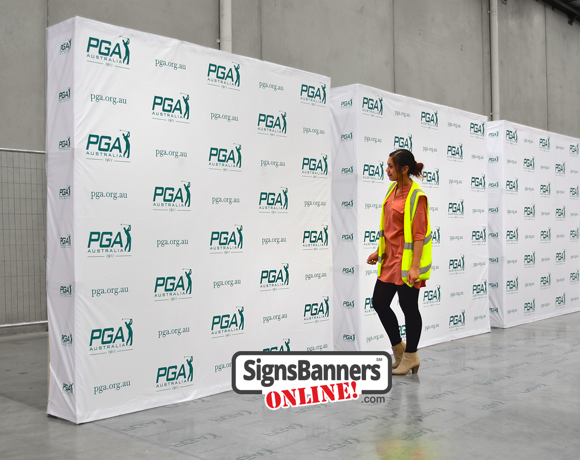 Woman in hi-vis vest beside tall white PGA Australia media wall pop-up displays in a warehouse.
