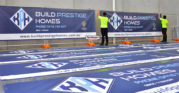 Two workers in hi-vis hanging printed Build Prestige Homes mesh banners with more banners laid out on the floor.